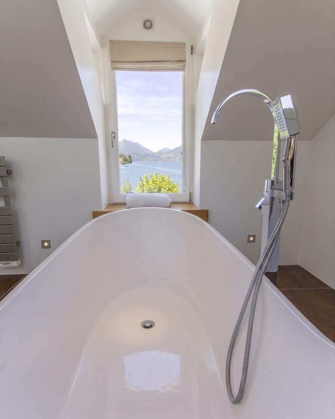 Freestanding bath beneath a dormer window framing a lake and mountain view