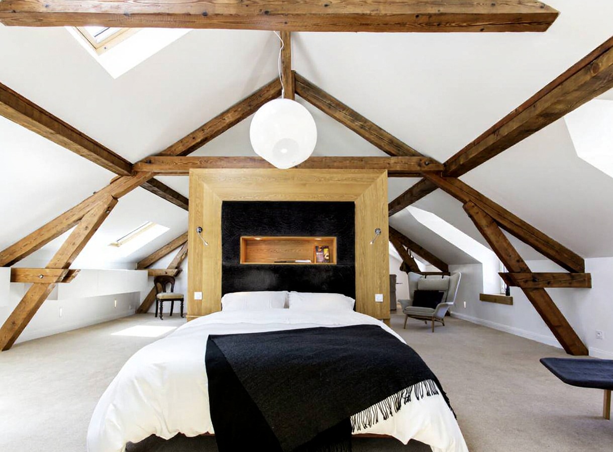 Attic bedroom with exposed timber trusses and dark headboard niche, precision carpentry under a vaulted ceiling.