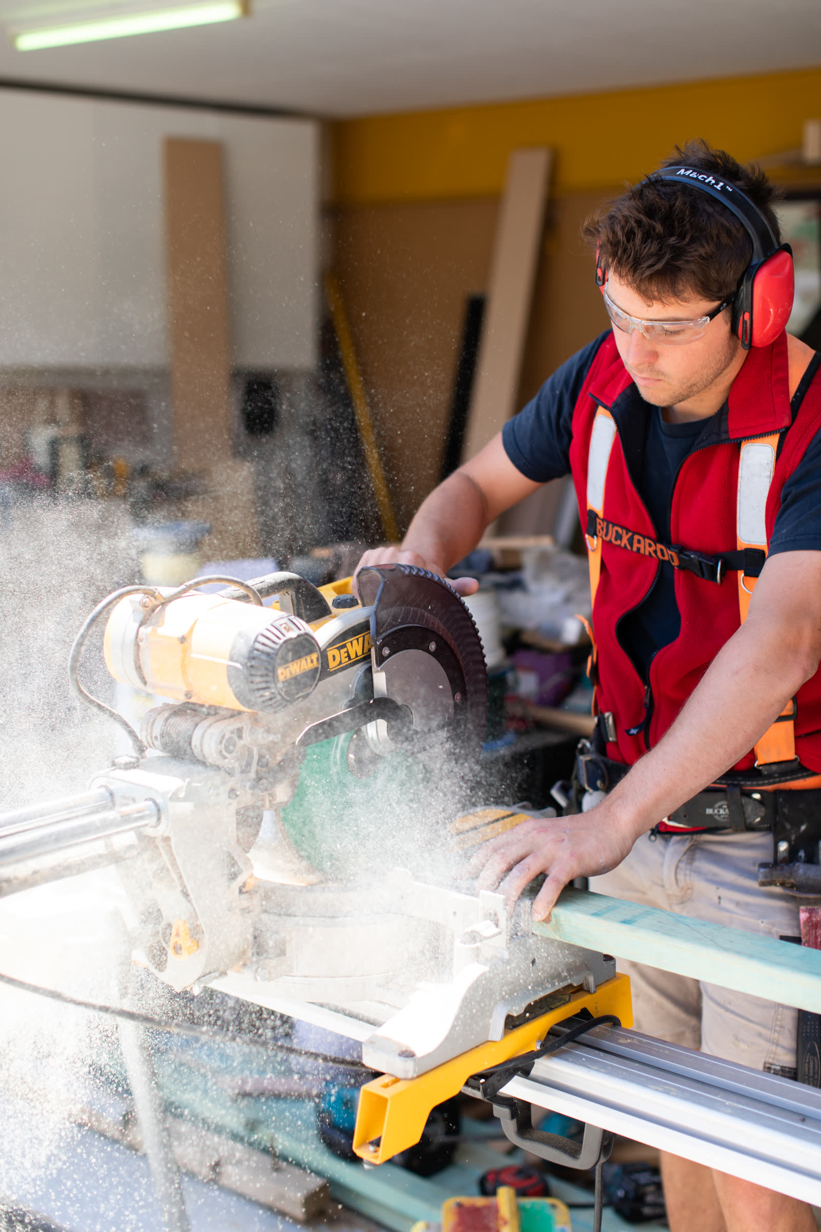 A Piper Moss carpenter cutting timber on a drop saw, wearing hearing protection, safety glasses and a hi-vis vest